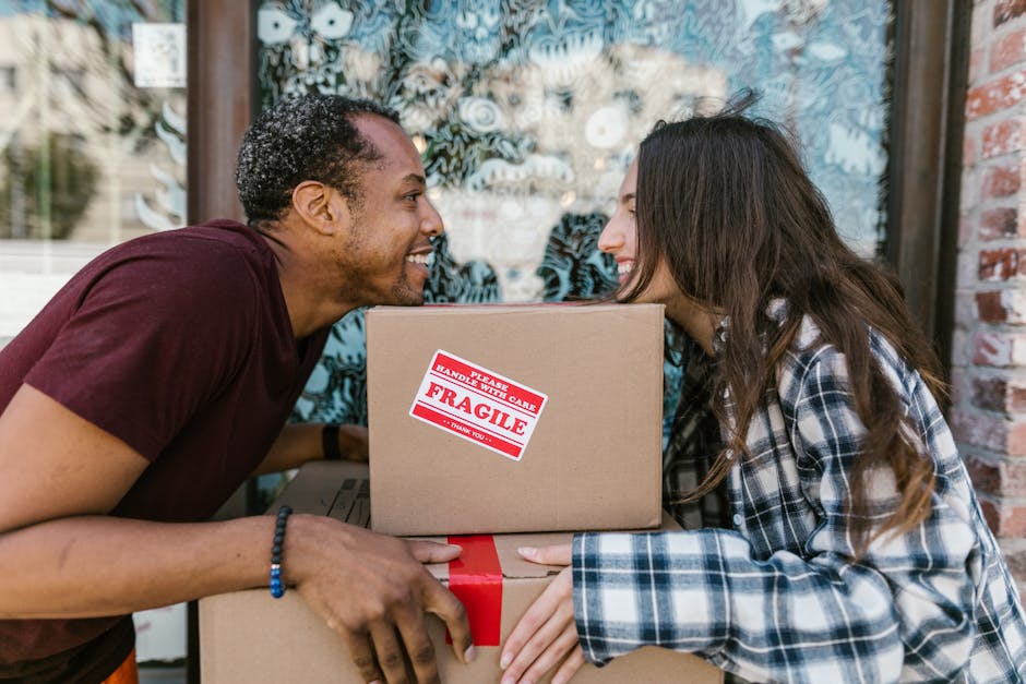 A man and a woman are engaged in a home relocation activity outside a building with a large glass window, preparing to load or unload cardboard boxes during a furniture transport process. The man, wea