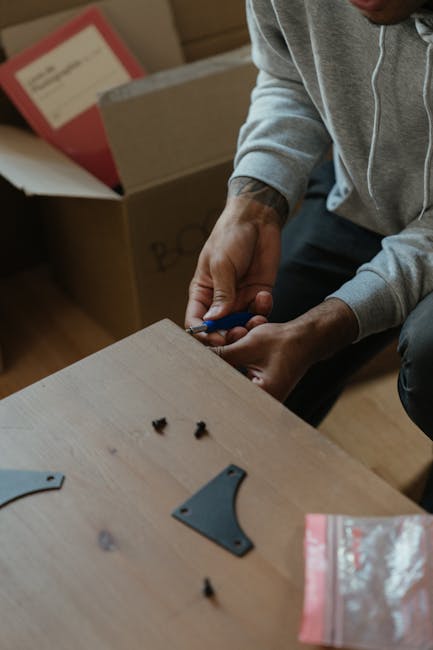 A person assembling furniture during a home relocation, using a screwdriver on a light wooden tabletop. The individual is dressed in a grey hoodie and has visible tattoos on their arm. Several small b