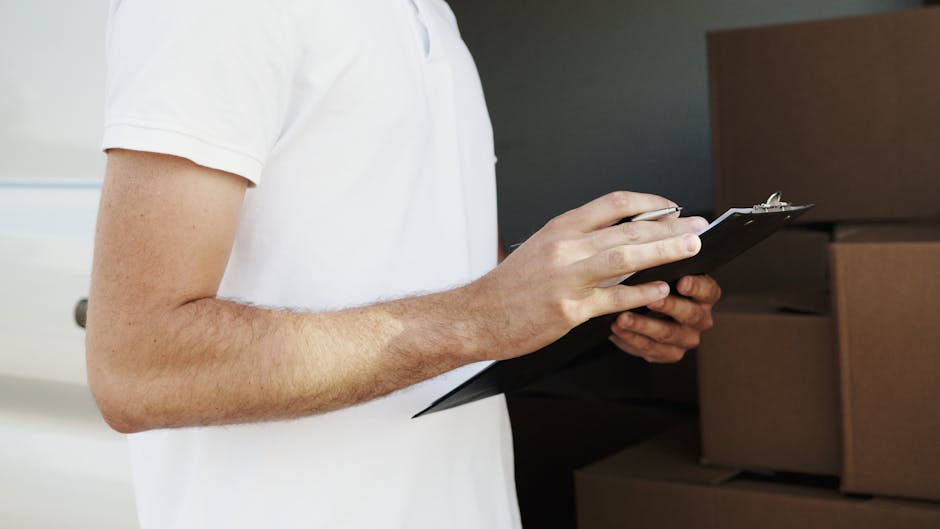 A male mover wearing a white shirt and jeans is holding a clipboard while discussing a house relocation with a female client inside a property. The woman, dressed in a blue and white checkered shirt,