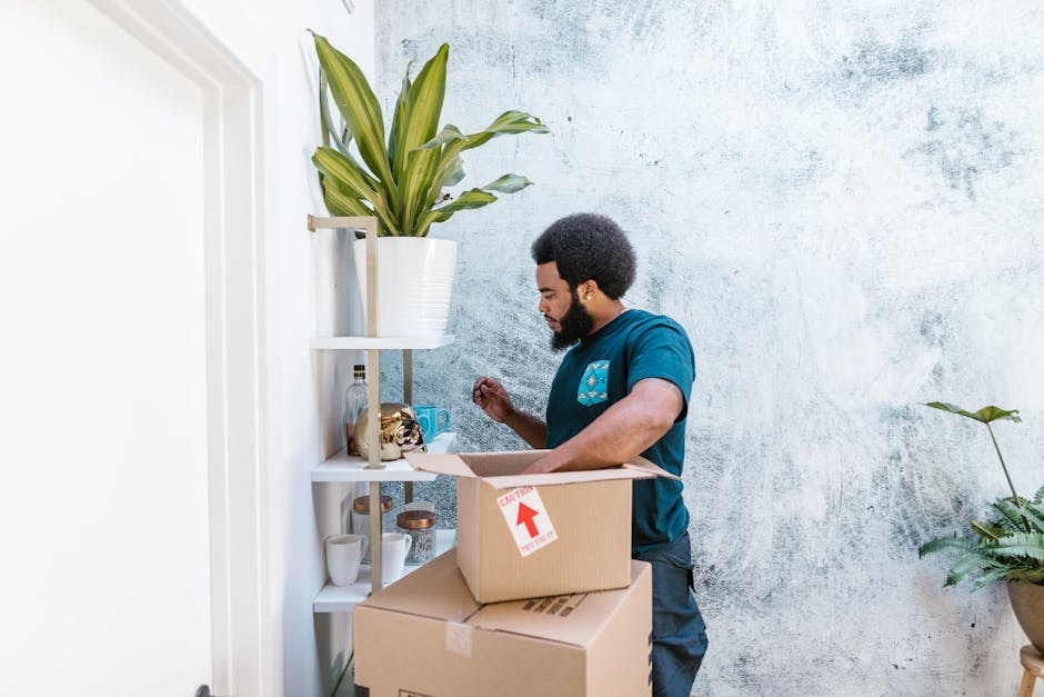 A woman and a young girl are seated on the wooden floor of a room during a home relocation process, surrounded by cardboard boxes of various sizes stacked behind them. The woman, dressed in a white t-