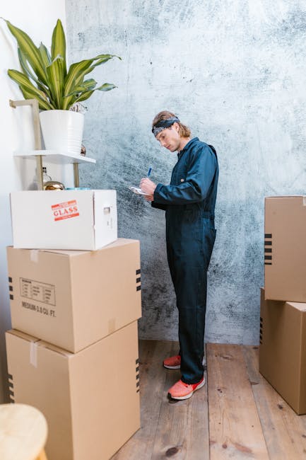A person dressed in a blue jumpsuit and wearing protective footwear stands in a room with a light blue textured wall, surrounded by stacked cardboard boxes and packaging materials. The boxes, some lab